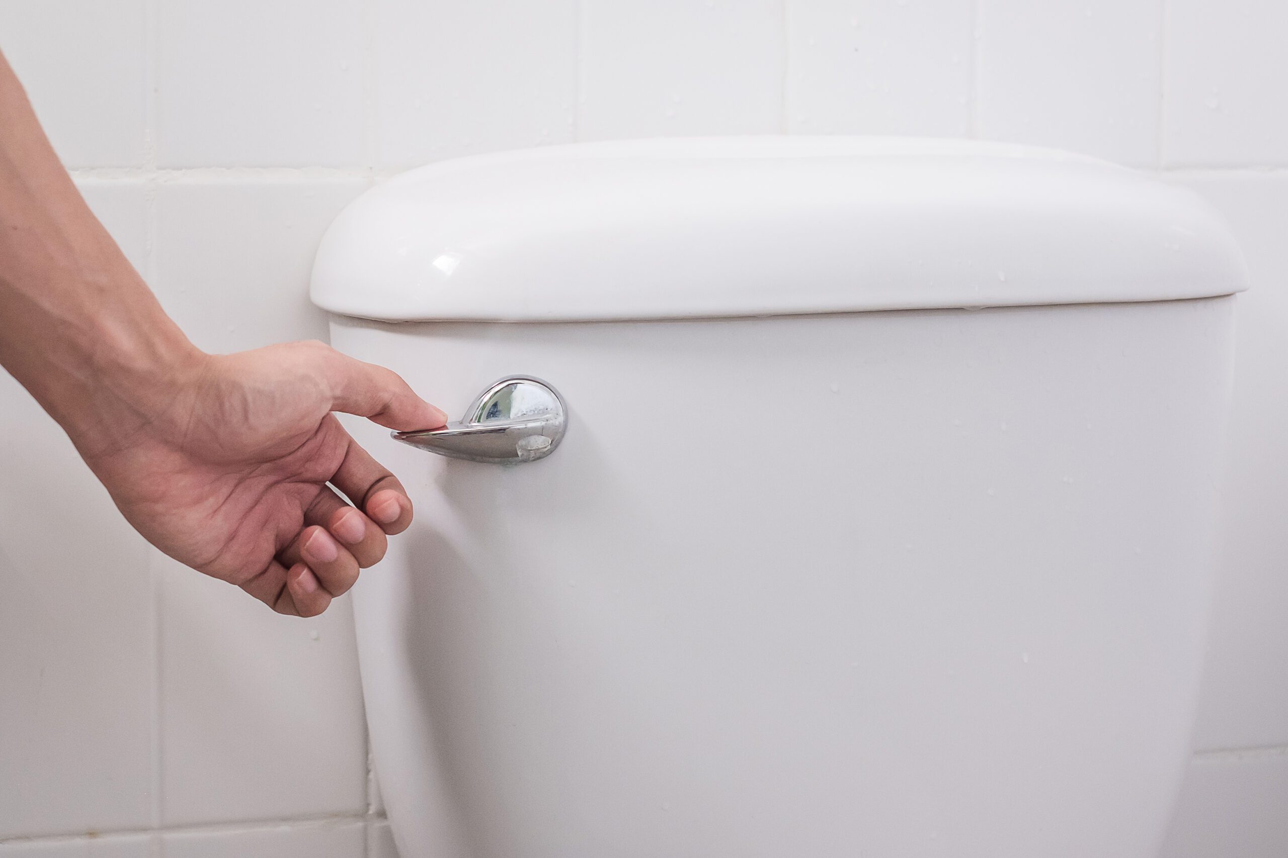 Hand flushing a modern white toilet using a chrome flush handle in a residential bathroom